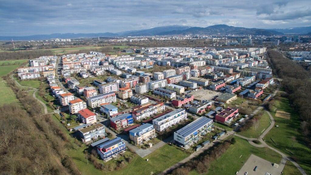 Luftaufnahme des Stadtteils Rieselfel mit vielen umweltfreundlichen Gebäuden, deren Dächer mitsamt ihrer blau-grauen PV-Anlagen zur Sonne ausgerichtet sind; im Hintergrund ist eine malerische Bergkette zu sehen