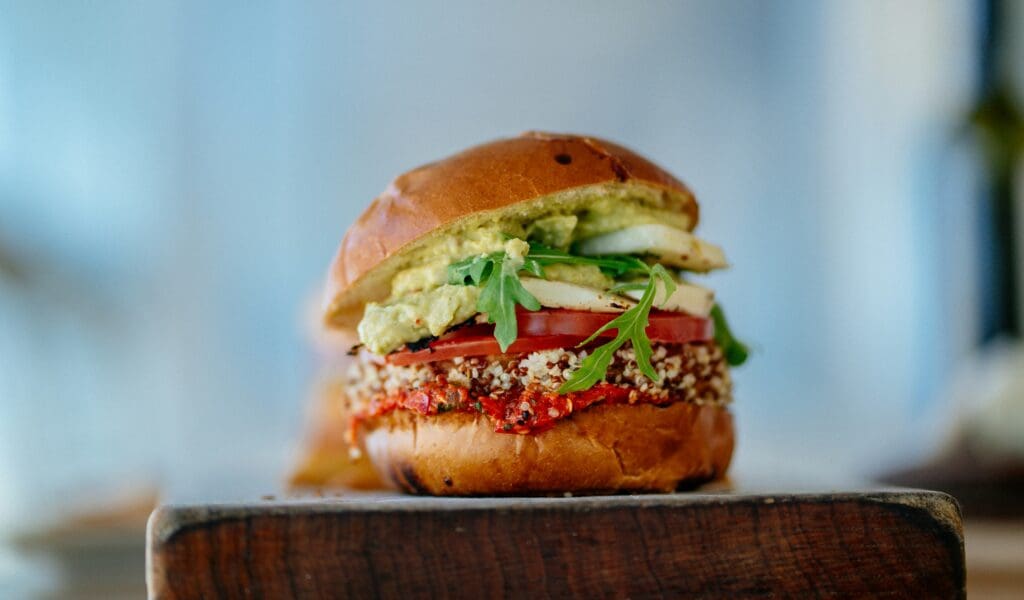 Veggie-Burger, präsentiert auf einem Holzbrett; auf dem Veggie-Patty liegen Schichten von Ruccola, Tomaten, gekrönt von einer Avocado-Sauce