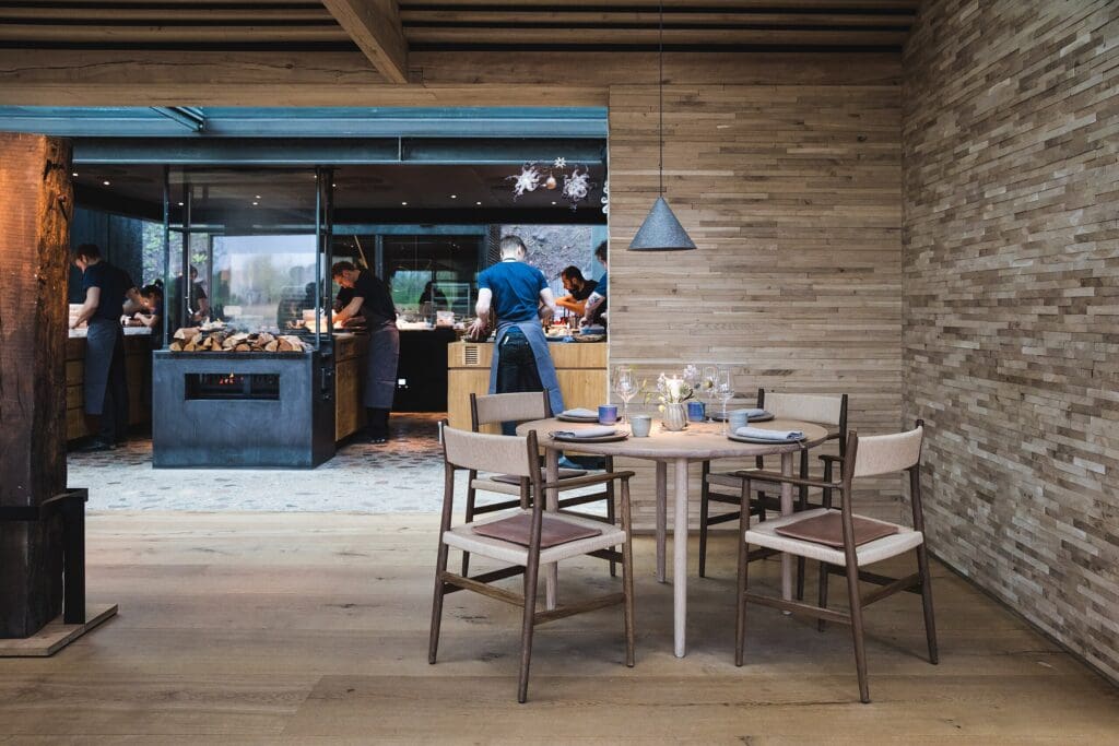 Im Michelin Restaurant "Noma" in Kopenhagen steht ein schön gedeckter Tisch im Vordergrund, während die offene Küche und das Servicepersonal den Hintergrund beleben.